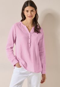 Camicia a maniche lunghe rosa con tessuto testurizzato, scollo a V con bottoni e polsini arrotolati. Abbinata a pantaloni bianchi per un look a contrasto.