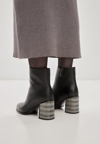 Botas de piel negras hasta el tobillo con un tacón en bloque gris texturizado y con patrón, y cremallera lateral, combinadas con una falda gris acanalada.
