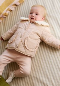 Veste matelassée rose rembourrée avec un col à volants et une poche, associée à un pantalon rose côtelé. Le fond présente un tissu rayé aux tons beige.