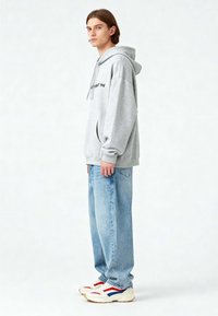 Felpa grigia con cappuccio e tasca davanti, con testo nero. Indossata con jeans baggy blu chiaro e sneakers bianche con dettagli rossi e blu.
