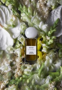 Flacon de parfum en verre ambre avec un bouchon rond blanc, étiqueté "Tiger by Her Side", entouré de fleurs blanches et vertes.