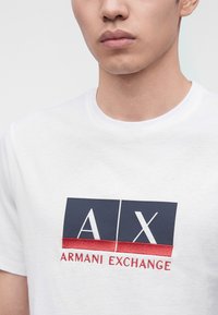 Άνδρας που φοράει λευκό μπλουζάκι με σήμα Armani Exchange σε ναυτικό μπλε και κόκκινο στο στήθος, περικομμένο κάτω από τη μύτη.