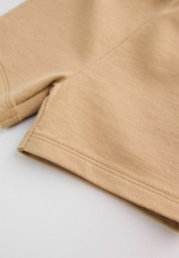 Gros plan sur un short en tissu beige montrant une double surpiqûre détaillée le long des ourlets et des coutures sur une surface plate blanche.