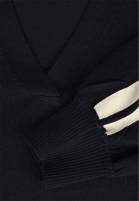 Pull noir en maille avec encolure en V profonde et poignets côtelés comportant deux bandes blanches horizontales sur la manche.