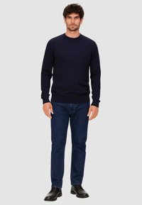 Sudadera de cuello redondo azul marino con puños y dobladillo acanalados, con un sutil logo "GUESS" en el pecho, combinada con jeans de mezclilla oscura y zapatos negros.