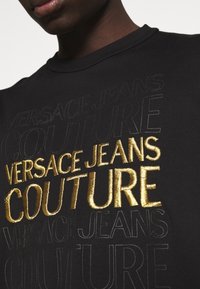 Schwarzes Baumwoll-T-Shirt mit einer Mischung aus schwarzem und goldenem Stickschriftzug, der "VERSACE JEANS COUTURE" in verschiedenen Schriftgrößen zeigt.