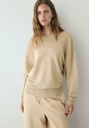 Giovane donna con capelli lunghi che indossa un maglione beige ampio e pantaloni abbinati, in piedi davanti a uno sfondo chiaro uniforme.