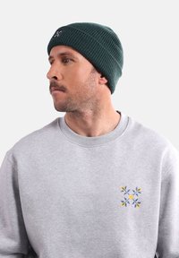 Bonnet en tricot vert avec un motif texturé. Porté avec un sweat-shirt gris clair arborant un design floral en jaune et bleu.