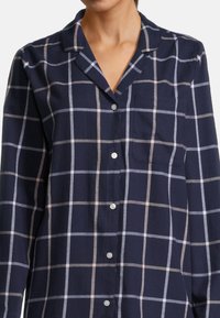 Donna che indossa una camicia blu navy con bottoni, caratterizzata da un grande motivo a scacchi bianco e rosa chiaro e una tasca sul petto.