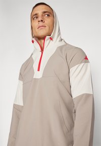 Lightweight, hooded sweatshirt with a half-zip closure. Features beige and white panels, red accents, and a textured fabric finish.