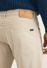 Chinos beige avec une texture lisse, arborant une étiquette en cuir sur la taille et une petite étiquette en tissu sur la poche arrière.