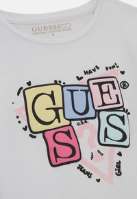 Valkoinen luomupuuvillapaita, jossa on värikkäitä lohkopainatuksia, joissa lukee "GUESS", "U.S.A" ja "JEANS", sekä leikkeitä.