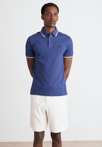 Marineblå polo skjorte med hvid kant, korte ærmer og et lille logo-patch, parret med lysebeige shorts. Glat bomuldsstof.