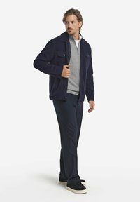 Marinblå knappad jacka i mjukt tyg, lagd över en grå dragkedjetröja, parat med mörka byxor och svarta sneakers.