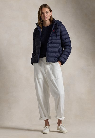 Noor naine, kellel on seljas tumesinine tikandiga jakk, must kampsun, lahtised valged püksid ja valged tossud, seisab käed taskutes.