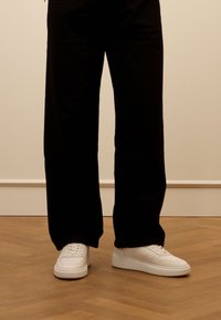 Weiße Sneaker mit einer glatten Lederoberfläche, runder Zehenpartie und dicker Sohle; kombiniert mit schwarzen weit geschnittenen Hosen auf einem Holzboden.