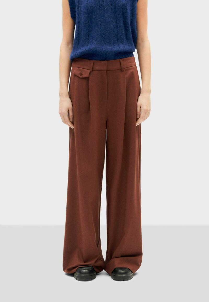 Pantaloni a gamba larga marroni con vita alta, realizzati in tessuto morbido; presentano un bottone anteriore, pieghe e tasche. Abbinati a un top in maglia blu navy.