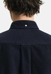 Marineblauwe corduroy hem met een klassieke kraag, textuurstof, een middenplooi en een witte knoop aan de achterkant.