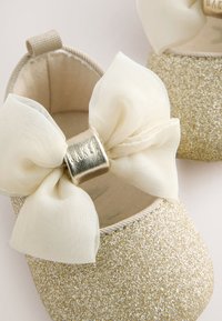 Chaussures bébé pailletées dorées avec grands nœuds en tissu crème et accents métalliques dorés au centre, doublure intérieure douce visible.