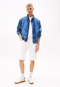 Jeansjakke med glidelås og ribbede mansjetter, hvit T-skjorte, hvite shorts og hvite sneakers med grønne detaljer. Enkelt bakgrunn.