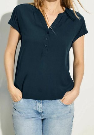 Woman wearing a short-sleeve navy blue buttoned blouse and light blue jeans with hands in pockets, standing against a plain background.