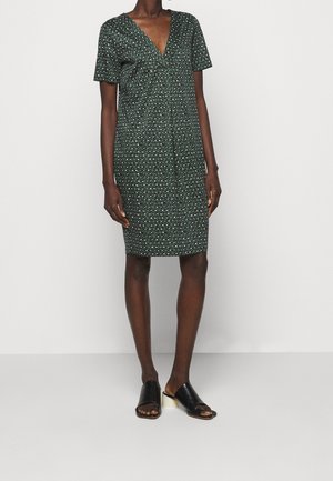 Kvinde iført en knælængde kjole med korte ærmer og et grønt geometrisk mønster samt sorte mule-sandaler med åben tå og hvide blokhæle.