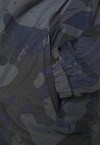 Vattentät kamouflagejacka i nyanser av mörkblått och grått. Har elastiska muddar, en dragkedjeficka och en slät yta med vattendroppar.