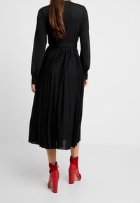 Czarna plisowana sukienka z długimi rękawami i paskiem w talii, połączona z czerwonymi botkami na obcasie z tłoczeniem węża. Gładka faktura, elegancka sylwetka.