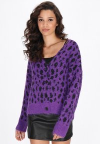 Paars fluffy cardigan met zwarte luipaardvlekken, knoopsluiting en lange mouwen. Draagt over een zwarte top en gecombineerd met een zwarte leren rok.