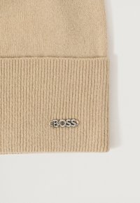 Beige kudumüts, millel on soonik tekstuur ja volditud äär. Esiküljel on hõbedane metallist logo.
