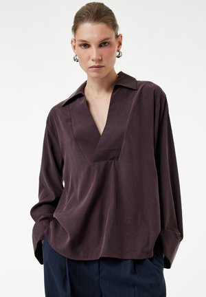 Donna con capelli tirati all'indietro indossa una blusa viola scuro a scollo a V con maniche oversize e pantaloni blu navy a righe sottili, con le mani in tasca.