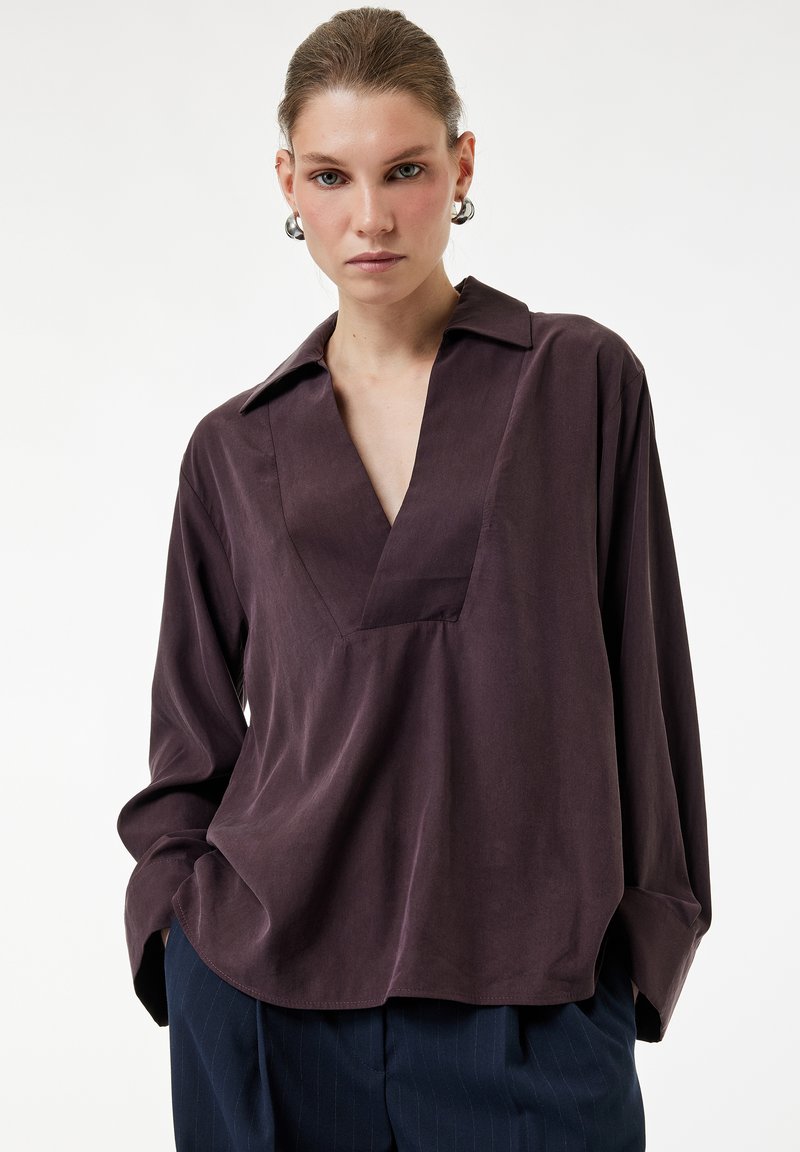 Mujer con el cabello peinado hacia atrás lleva una blusa morada oscura con cuello en V y mangas sobredimensionadas, y pantalones azul marino con pinzas, con las manos en los bolsillos.