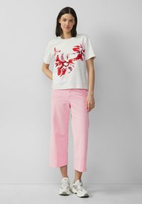 Weißes Baumwoll-T-Shirt mit rotem Blumenmuster, kombiniert mit hellrosa, verkürzten Hosen. Die Schuhe sind graue Sneakers mit weißen Akzenten.