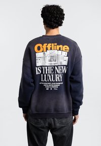 Námořnický svetr se značným oranžovým a bílým textem na zadní straně. Text obsahuje "Offline" a "JE NOVÝM LUXUSEM." Měkká látka a uvolněný střih.