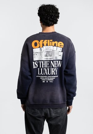 Sudadera navy con texto en naranja y blanco en la parte posterior. El texto incluye "Offline" y "ES EL NUEVO LUJO." Tejido suave y corte relajado.