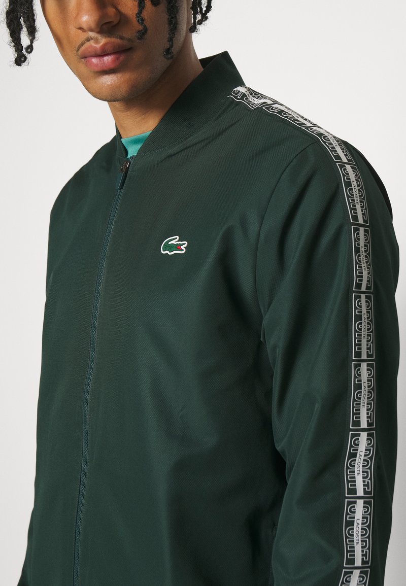 Bómber de color verde oscuro hecho de material texturizado. Presenta un cierre de cremallera y cinta de logo en contraste a lo largo de los hombros. Logotipo de cocodrilo de Lacoste visible.
