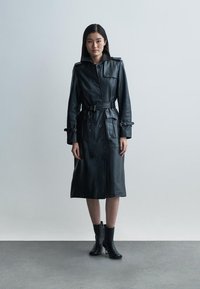 Black leather trench coat with a structured collar, belted waist, and side pockets. Smooth texture with buttons and adjustable cuff straps.