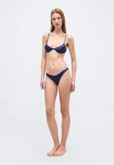 Νεαρή γυναίκα που στέκεται ξυπόλητη φορώντας μπλε σκούρο μπικίνι με λεπτές τιράντες, μπροστά σε απλό ανοιχτό φόντο.