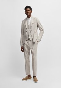 Helle, strukturierte Anzug mit einem Zwei-Knopf-Blazer, passender Weste und schmalen Hosen. Getragen mit einem weißen Hemd und hellbraunen Slippern.