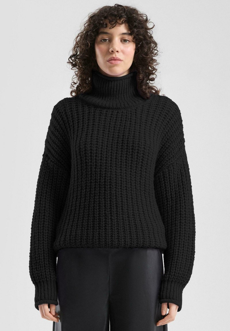 Zwarte gebreide trui met een hoge col, wijde mouwen en een gestructureerd patroon. De stof oogt dik en knus. Gecombineerd met een donkere broek.