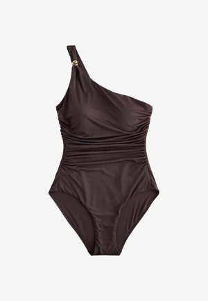 Fekete egyrészes, egyik vállat szabadon hagyó fürdőruhát viselő nő áll egy homokos tengerparton egy nagy szikla mellett, jobb csuklóján több arany karkötővel.