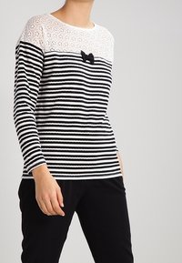 Raidallinen pitkähihainen paita, jossa on pitsiolkaimet, musta-valkoiset raidat ja pieni musta nauha yksityiskohtana edessä.