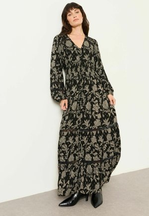 Frau trägt ein langes schwarzes Blumenkleid mit langen Ärmeln und schwarzen spitzen Stiefeletten, steht an einer weißen Wand.
