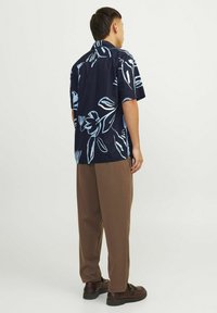 Camicia a maniche corte con sfondo blu navy e motivi floreali azzurri, abbinata a pantaloni marroni e scarpe scure.