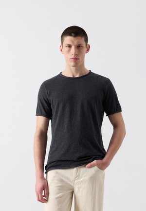 Noor mees lühikeste tumedate juustega, kes kannab tumedat halli lühikese varrukaga särki ja helebeežid pükse, seistes lihtsa heleda tausta ees.