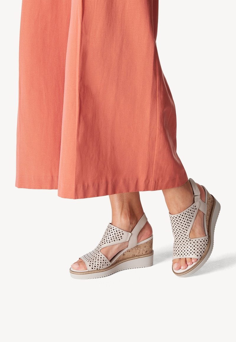 Sandales compensées beiges à design perforé portées avec un pantalon corail à jambes larges, dévoilant chevilles et pieds sur un fond uni.