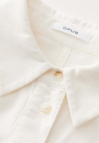 Cream-colored cotton shirt featuring a ribbed texture, a classic collar, and two buttons at the front. Label reads "OPUS."