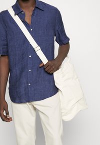 Navyblaues Leinenhemd mit kurzen Ärmeln und Knöpfen, kombiniert mit crèmefarbenen Hosen. Das Model trägt eine weiße Canvas-Tasche mit einem Riemen.