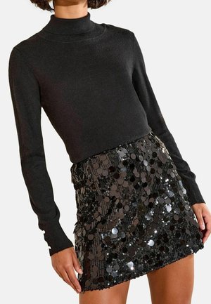 Mujer con un suéter negro de cuello alto y manga larga y una mini falda negra con lentejuelas, de pie frente a un fondo liso.