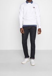 Felpa bianca con scollatura rotonda e logo ricamato, indossata sopra una camicia blu con colletto, abbinata a jeans scuri e sneakers bianche.